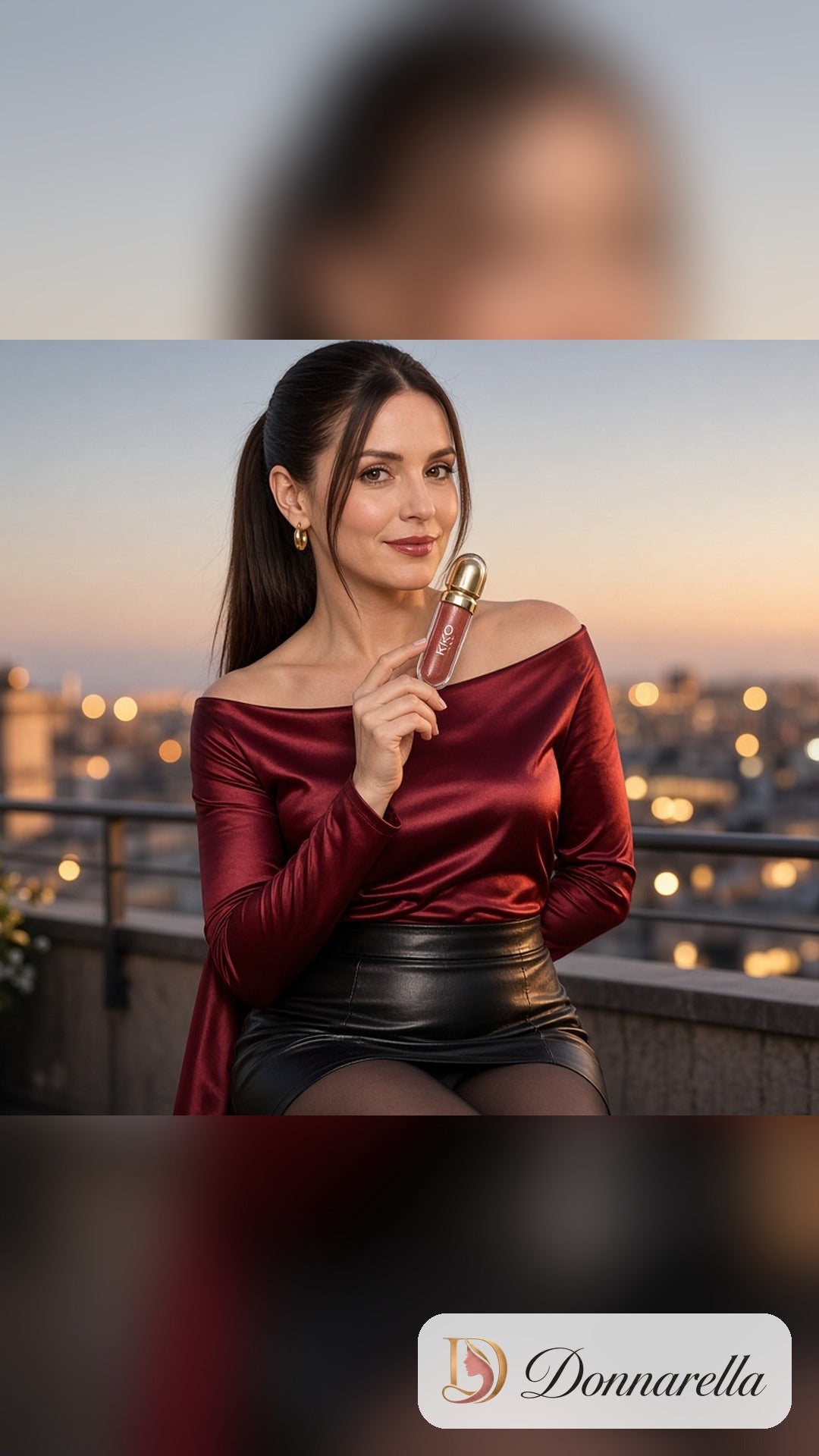 Nossa modelo com rabo de cavalo alto, sem óculos, de blusa ombro a ombro de cetim vinho e saia preta, segurando o Gloss Labial KIKO MILANO em um terraço ao pôr do sol.
