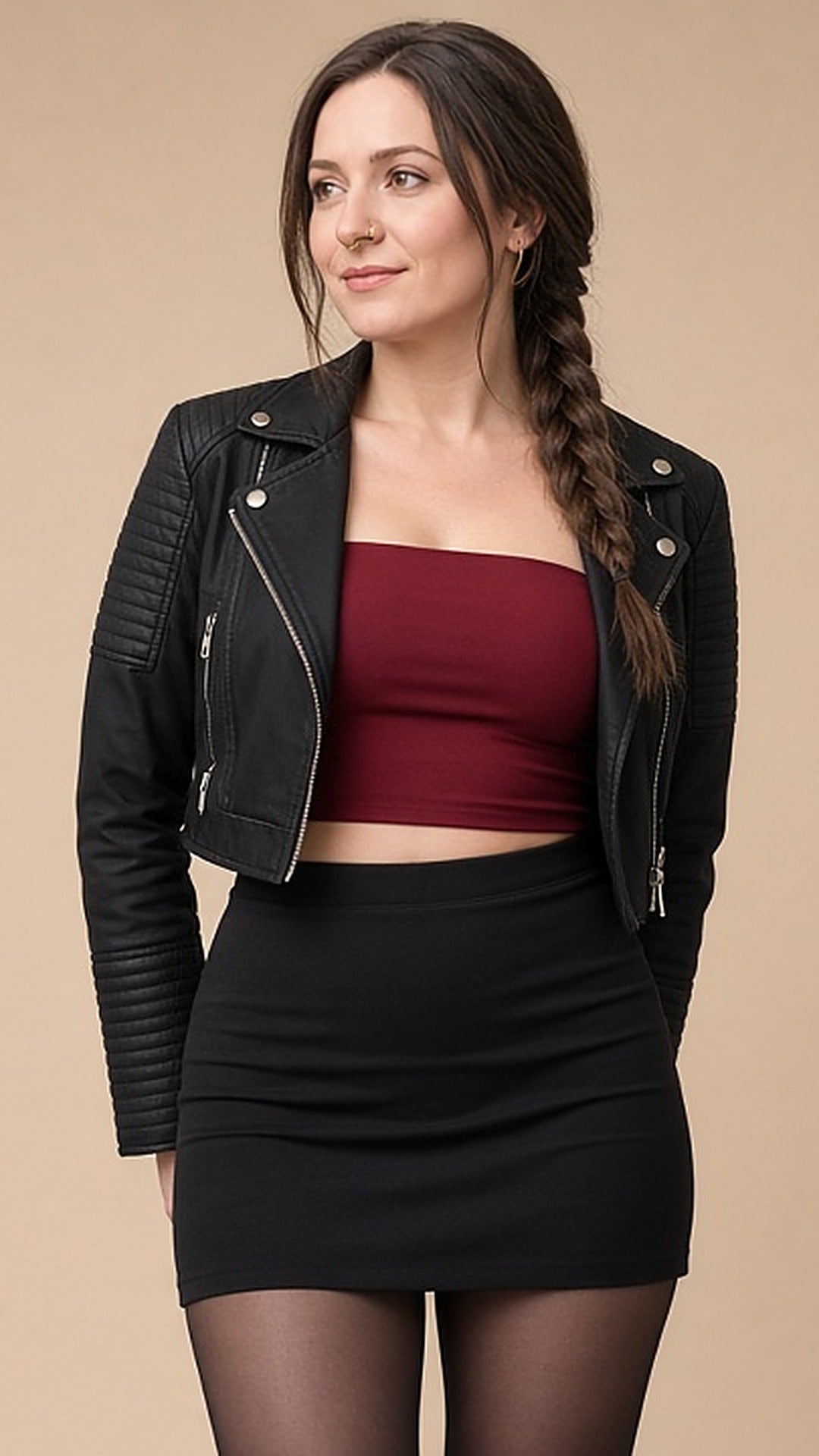 Modelo Donnarella sem óculos, cabelo em trança lateral, usando piercing fake de nariz argola dourada formato D, jaqueta estilo motoqueira preta, top tomara que caia vinho, minissaia preta e meia-calça
